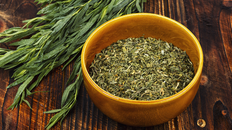 Stems of fresh, green tarragon plants lay beside an orange bowl of dried tarragon herbs