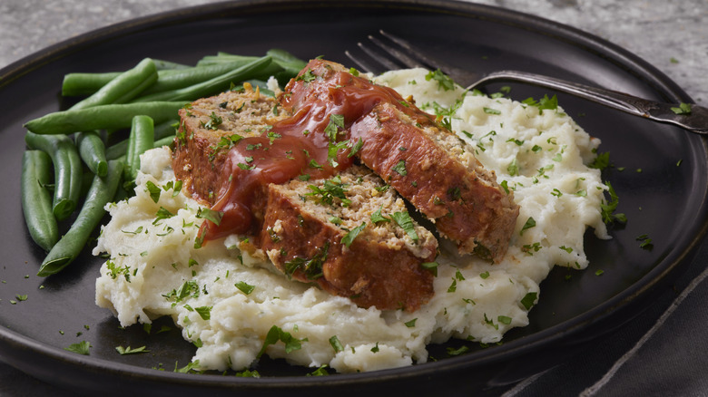 meatloaf covered in sauce with mashed potatoes, herbs, and green beans on a plate with fork