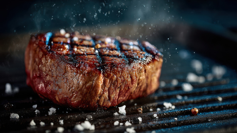 A perfectly grilled steak sits on a hot grill pan, glistening with rendered fat and sprinkled with coarse salt