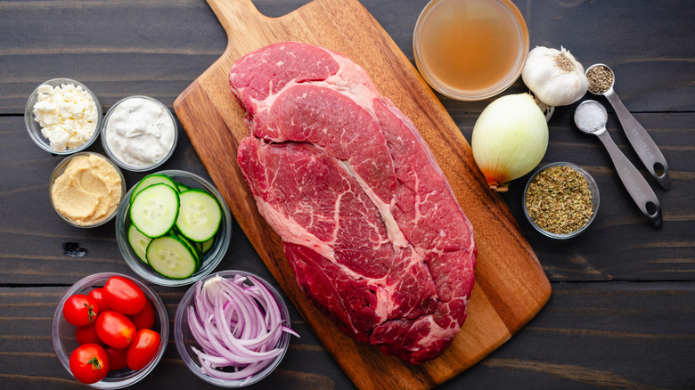 Chuck roast on a wooden cutting board, with beef broth, onion, oregano, grape tomatoes, sliced cucumber, and more in ramekins