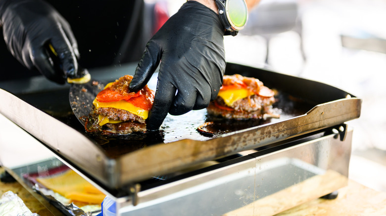 Chef preparing smash burgers on a flat top grill that are topped with tomatoes and cheese