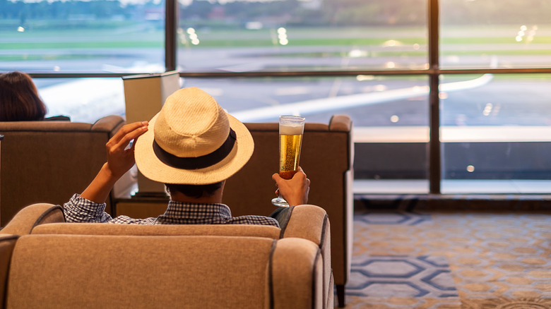 A man in a wide-brimmed hat holding a champagne glass and looking out an airport window