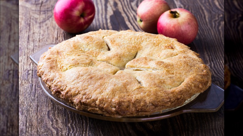 Whole Amish Apple Schnitz Pie sitting on plate on wooden table with three whole fresh apples behind it