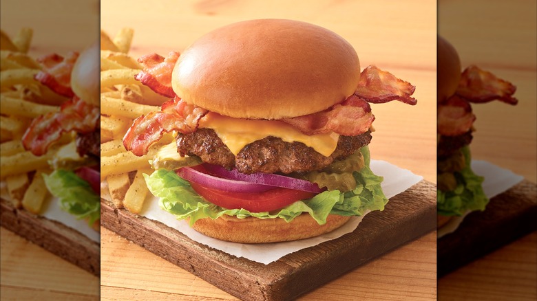 A bacon cheesburger with tomato, lettuce, and onion with fries on a wooden cutting board.