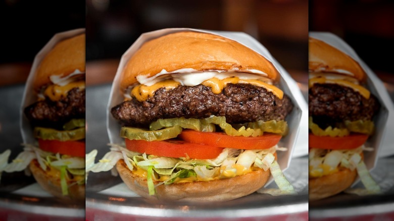 A thick burger from Fuddruckers with mayo, mustard, pickles, tomato, and lettuce on metal tray.