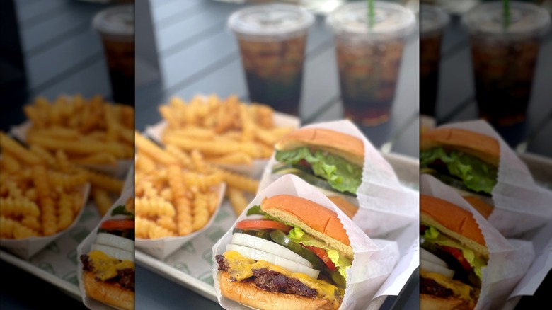 Two cheeseburgers in wrappers arranged on a silver tray next to sides of crinkle cut fries and two sodas.
