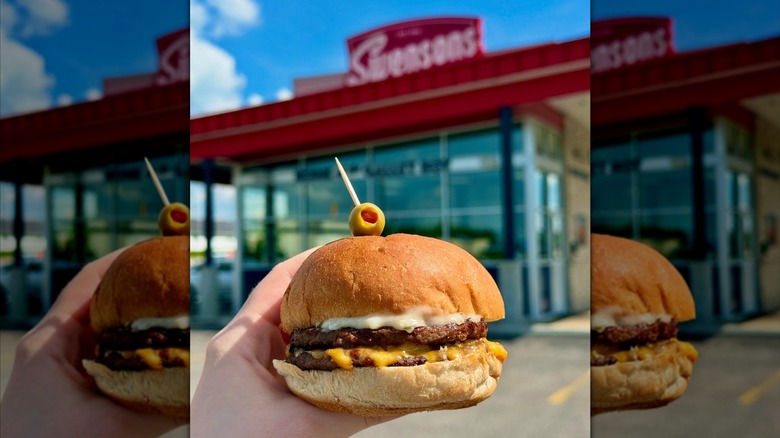 Someone holding a double cheeburger with olive on top in front of Swensons location.