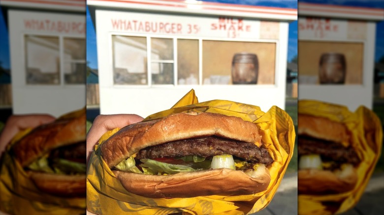 Someone holding a paper-wrapped cheeseburger outdoors in front of Whataburger's original location.