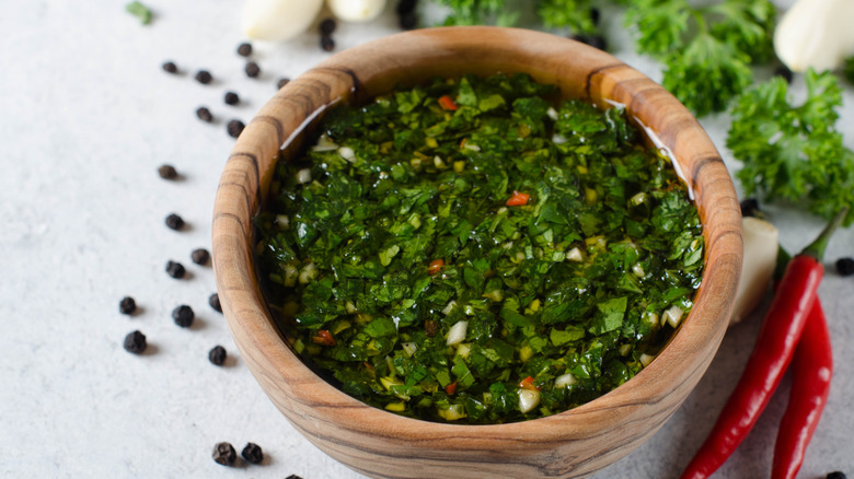 A wooden bowl of homemade chimichurri sauce surrounded by fresh ingredients