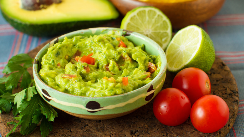 A bowl of guacamole surrounded by fresh tomatoes, lime, avocado, and cilantro