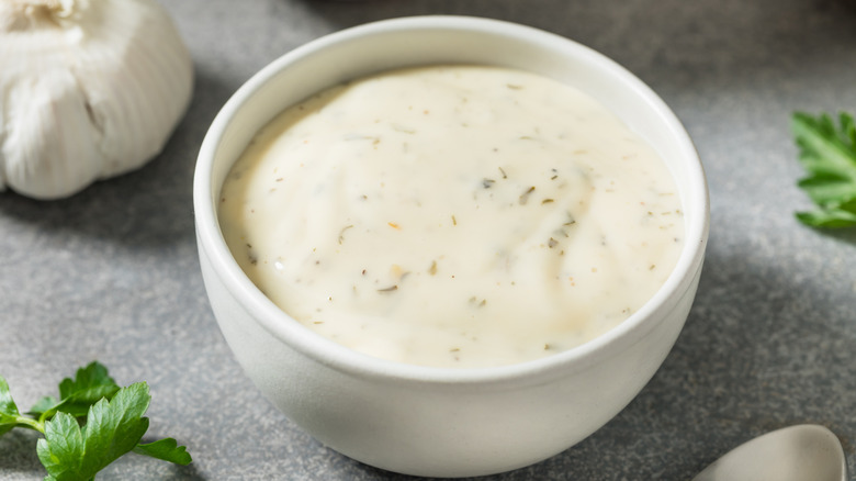 A ceramic ramekin full of homemade ranch dressing on a countertop surrounded by fresh parsley and garlic