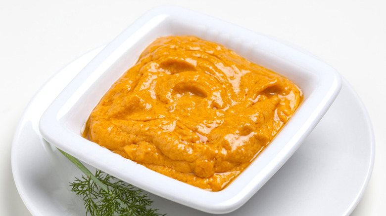 A white dish of romesco sauce on a saucer garnished with a fresh herb sprig