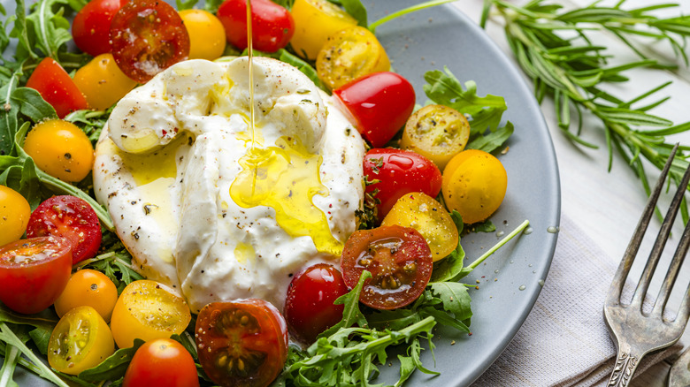 Olive oil being drizzled over a salad of burrata, cherry tomatoes, and arugula