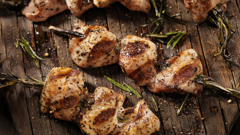 Barbecued chicken kabobs on rosemary stems on wooden board surrounded by rosemary leaves