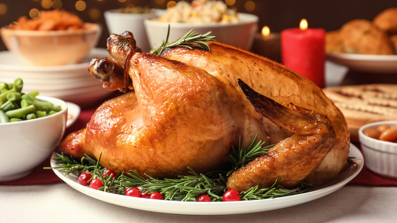 A roasted turkey surrounded by rosemary and red berries on white plate in front of other white bowls of holiday side dishes.