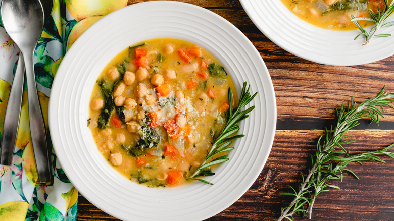 Vegetable chickpea soup in a white bowl with fresh rosemary on wooden table