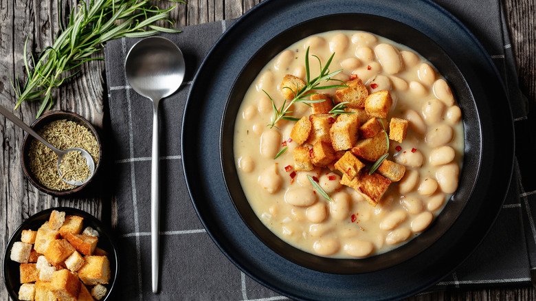 White beans in a black bowl topped fresh rosemary and croutons
