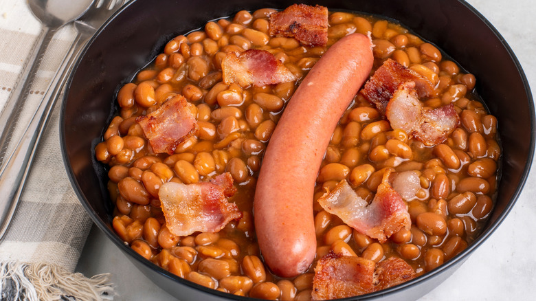 A skillet of baked beans with bacon and a hot dog in the middle