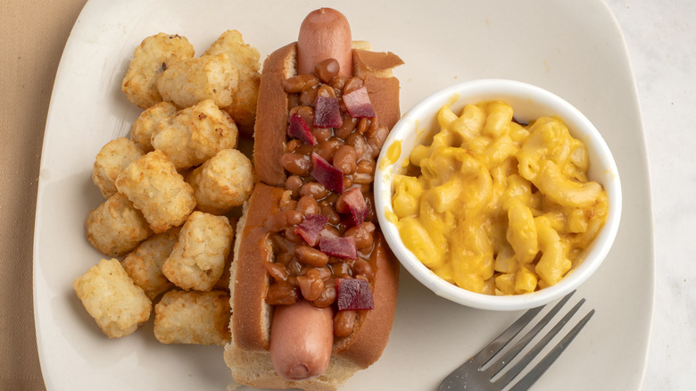 Baked beans on a chili dog on a white plate with macaroni and cheese and tator tots and a fork