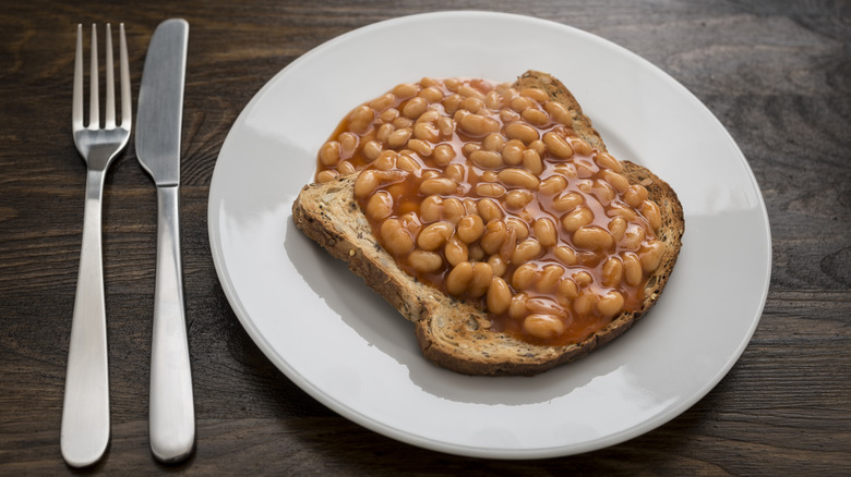 Baked beans over toast on white plate on top of dark table with fork and knife on the side