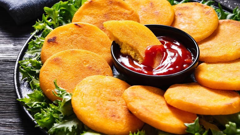 A small ketchup ramekin positioned in the middle of a circle of hot water cornbread patties resting on a bed of lettuce