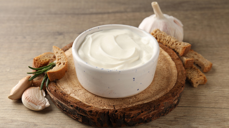 Ramelin of white sauce, bread strips, and garlic cloves on wooden cutting board