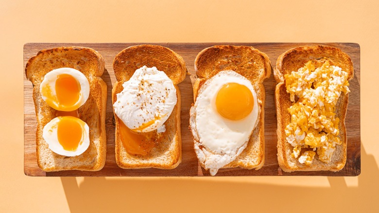 Four pieces of toast on a cutting board topped with boiled eggs, poached eggs, fried eggs, and scrambled eggs