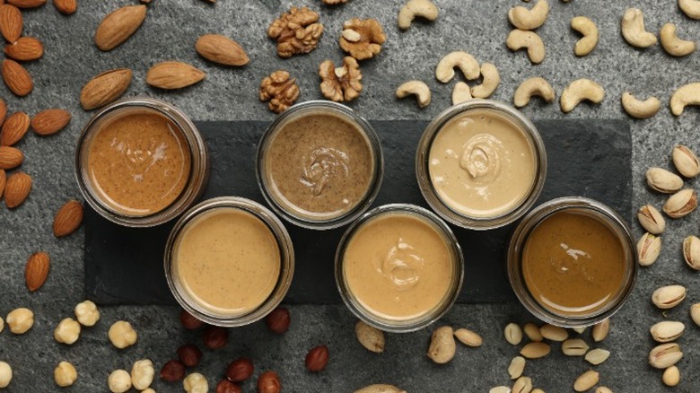 top view of six jars of assorted nut butters surrounded by assorted whole nuts on a gray background