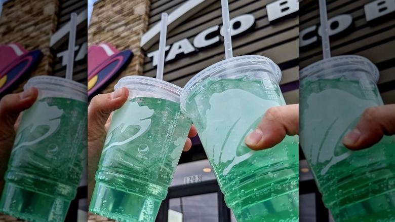 two hands hold clear, plastic cups of Baja Blast in front of a Taco Bell storefront