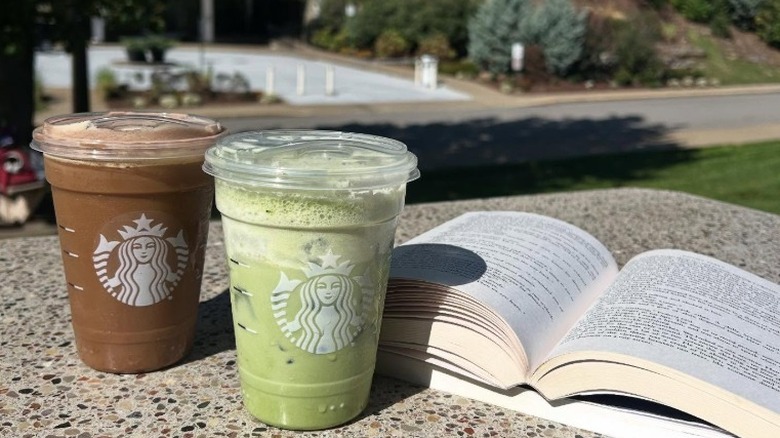 An Iced Banana Cream Protein Matcha & Iced Chocolate Cream Protein Cold Brew from Starbucks sit next to an open book