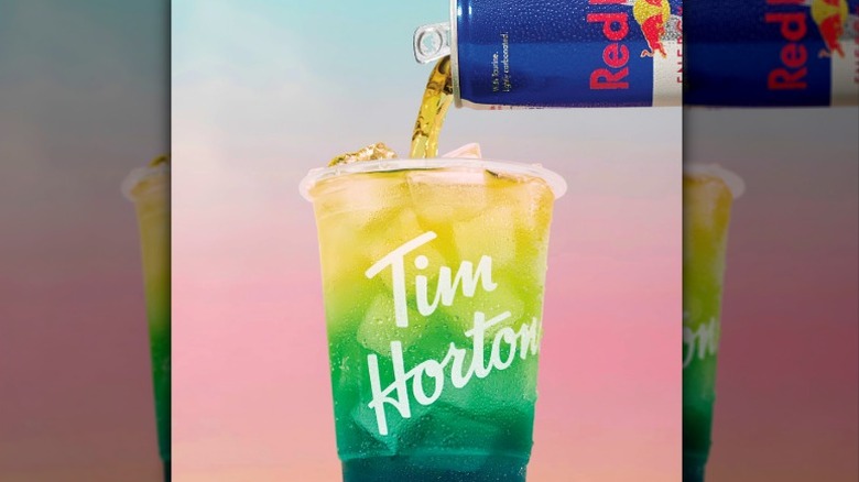 Liquid pours from a can of Red Bull into a Tim Hortons cup filled with fizzy soda and ice cubes