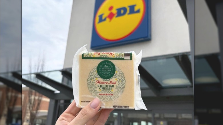 Someone holding a hunk of cheddar cheese in front of Lidl.