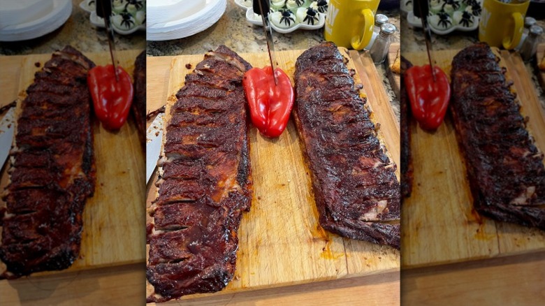 two racks of ribs on a wooden board surrounding a whole red pepper with a knife stuck in it