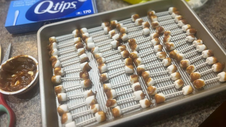 pan of mini marshmallows dipped in caramel sauce stuck to each end of lollipop sticks posed in front of a box of Q-tips