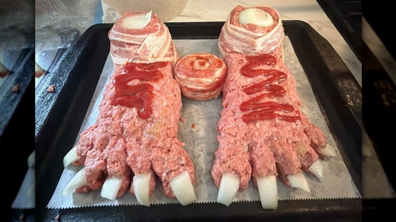 two uncooked meatloaves shaped like feet and decorated with sliced onions, bacon strips, and ketchup sitting in a paper-lined pan