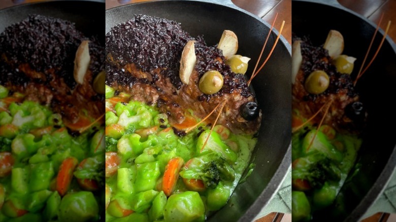 rat-shaped meatloaf decorated with potatoes, olives, spaghetti, and black rice in a pan alongside mixed vegetables dyed with green food coloring