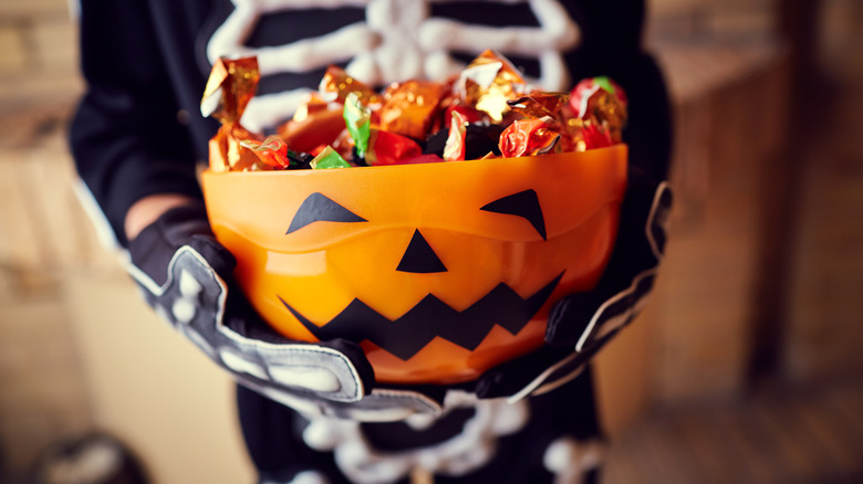 A boy in a skeleton costume holding an orange bowl with candy inside.