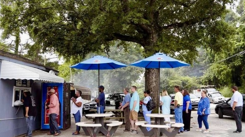 A group of customers lined up in front of B's Barbecue among white picnic tables with blue umbrellas