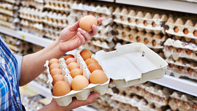 A person holding a carton of eggs in one hand and an egg from that carton in another standing in front of a display of egg cartons