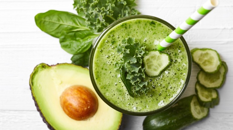 Top view of a glass of green juice next to sliced cucumber, a halved avocado, and leafy greens