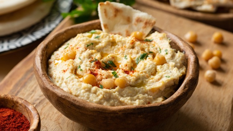 A wooden bowl with hummus topped with chickpeas, paprika, and pita bread sits on a wooden cutting board