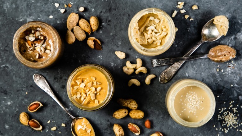 4 jars of nut butters on a black service surrounded by spoons of nut butter and raw nuts