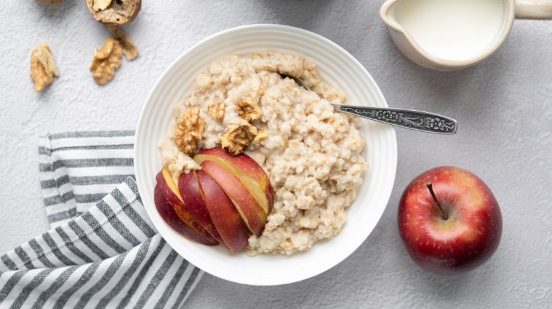 A bowl of oatmeal with sliced apples and walnuts on a white surface next to a whole apple, a cup of milk, and a striped cloth