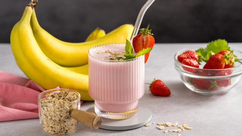 a strawberry banana smoothie on a countertop next to a bowl of strawberries, a measuring cup with oats, and a bunch of bananas on a pink towel