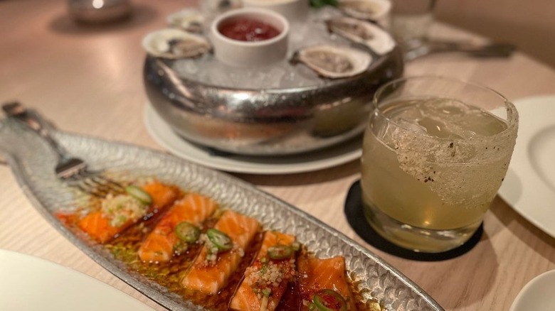 A hammered silver platter with citrus cured salmon next to a cocktail and oysters on an ice bed