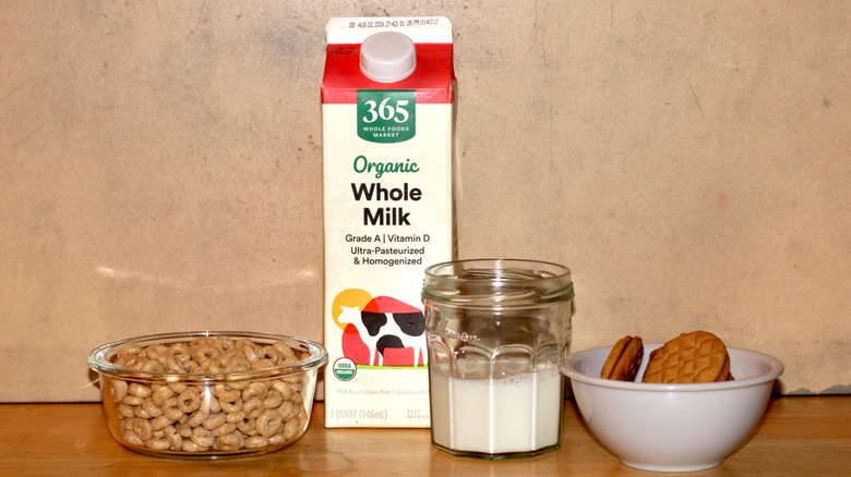 A 365 brand carton of organic whole milk beside a glass of milk, a bowl of cereal, and some cookies