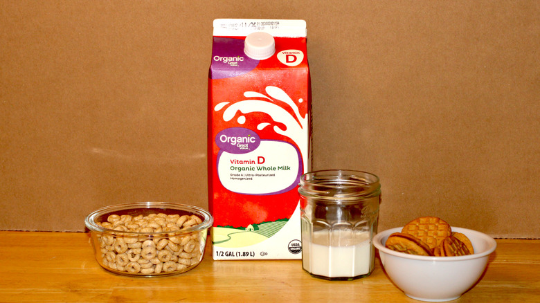 A Great Value brand carton of organic whole milk beside a glass of milk, a bowl of cereal, and some cookies