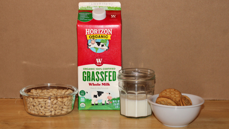 A Horizon brand carton of organic grass fed whole milk beside a glass of milk, a bowl of cereal, and some cookies