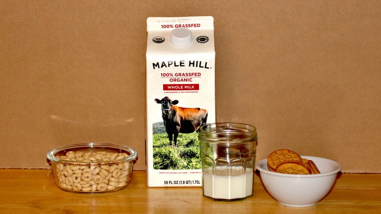 A Maple Hill brand carton of organic grass fed whole milk beside a glass of milk, a bowl of cereal, and some cookies