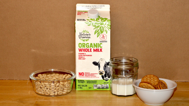 A Nature's Promise brand carton of organic whole milk beside a glass of milk, a bowl of cereal, and some cookies